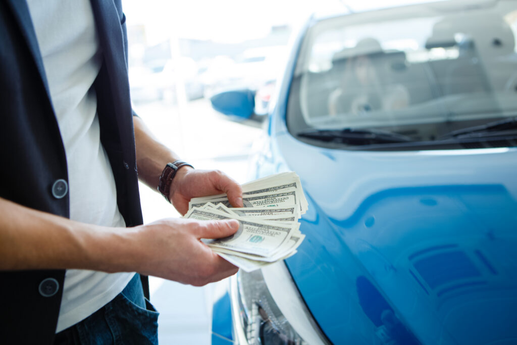 A person holding money with a blue car in the background.